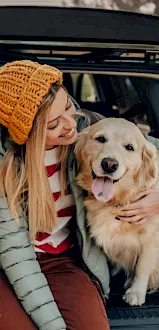 A woman in a knit orange beanie cuddles a golden retriever while sitting in a car trunk.